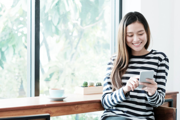 young-asian-woman-using-smart-phone-while-sitting-by-window-cafe-background_7190-874